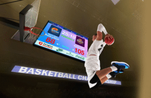 Jugador de baloncesto realizando un mate frente a un gran marcador LED digital.