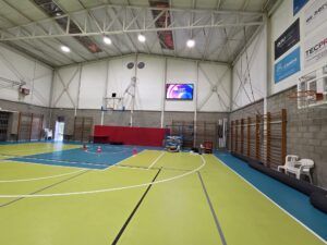 Interior de un pabellón deportivo con canasta y un gran videomarcador LED en la pared frontal.